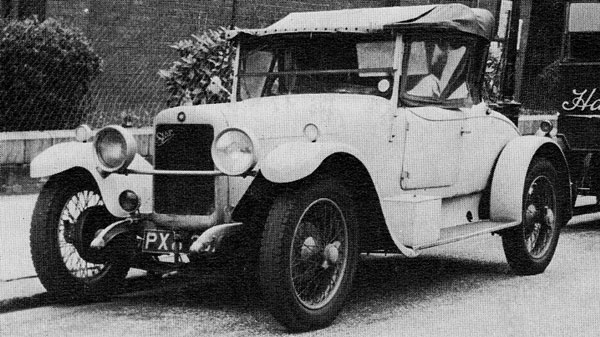 A vintage car is parked by the side of the road. The car has a classic design, featuring a rounded front and a prominent grille. Its body is predominantly white, with black accents on the wheels and parts of the trim. The vehicle has a convertible soft top, partially raised, and displays a number plate that begins with "PX." In the background, there are some bushes and a section of a building, adding context to the urban environment. The overall look conveys a nostalgic charm typical of early 20th-century automobiles.