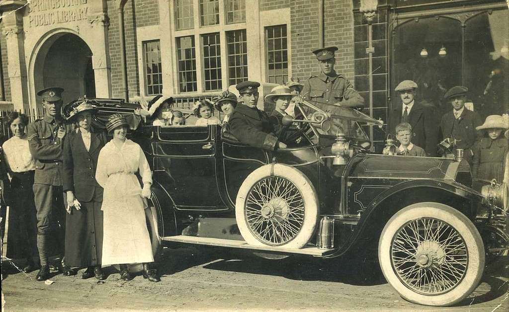 The photo features a large group of people standing in front of a historic building, identified as a public library. In the foreground, there is a vintage car with a glossy black finish and large, white tires. Several individuals are seated in the car, while others stand around it. 

To the left, a soldier in uniform poses beside two women, one wearing a hat and the other dressed in a blouse and skirt. Next to them, a woman in a long white dress stands with her hands on her hips. Other figures include children and adults wearing various vintage clothing styles, including hats. The background shows the library’s ornate brick facade and large windows. The overall atmosphere suggests a historical moment, perhaps a celebration or community gathering.