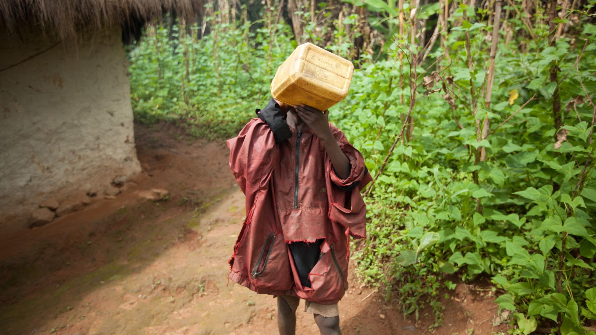 A child stands on a dirt path surrounded by lush greenery, carrying a yellow water container on their head. The child is dressed in a tattered red jacket that hangs loosely, and they have their hands positioned around the container. Behind them, a small thatched-roof structure can be seen, partially obscured by the dense vegetation. The atmosphere appears humid, suggesting a rural setting rich in plant life.
