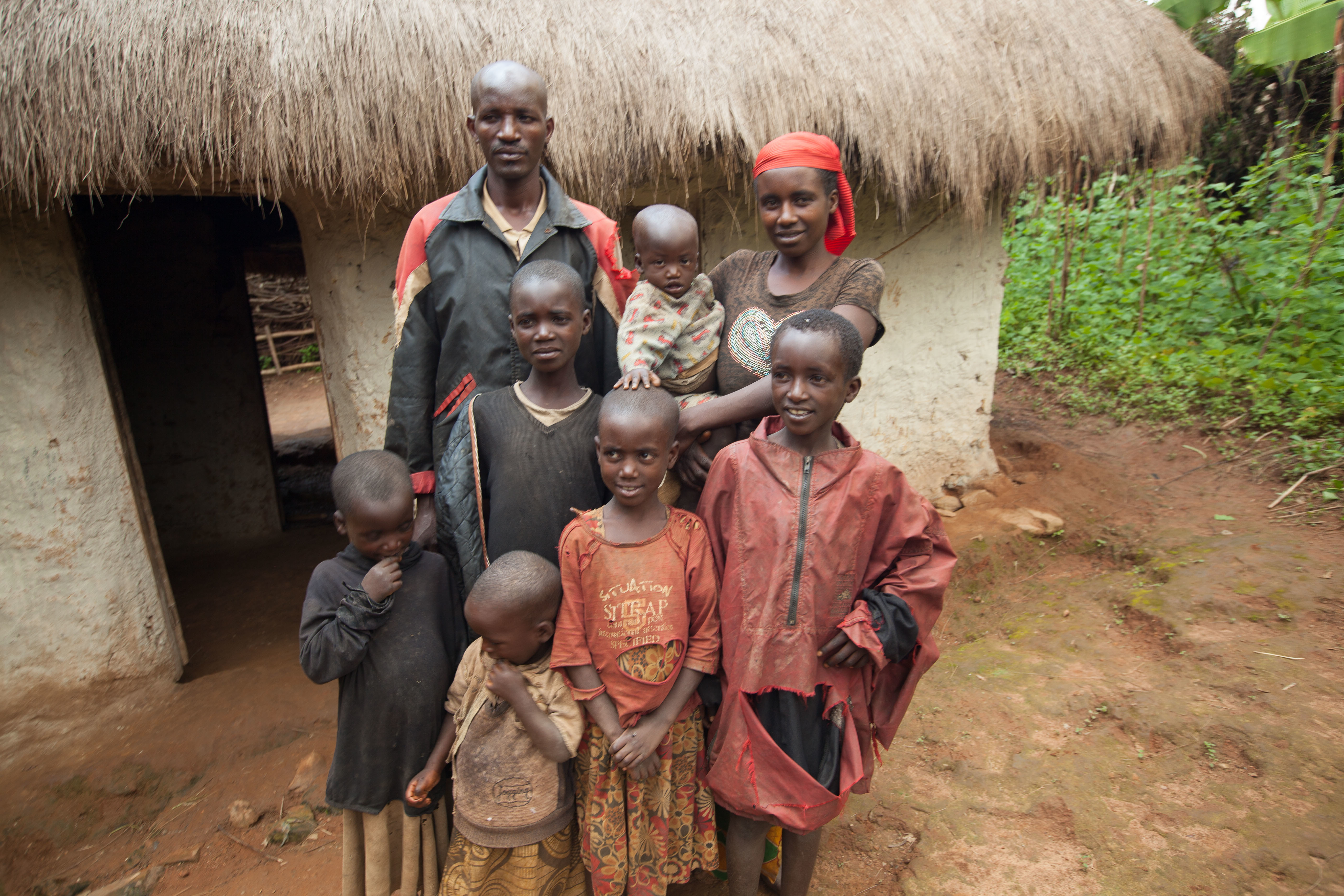 A family stands outside a small, rustic house with a thatched roof. The father, wearing a black and red jacket, stands at the back. Next to him is the mother, holding a baby. In front of them are five children of varying ages, all wearing simple, somewhat worn clothing. The children display a mix of expressions, from shy smiles to more serious looks. Behind them, the landscape features lush green foliage and dirt ground, giving a glimpse of their rural setting. The atmosphere appears warm and familial.