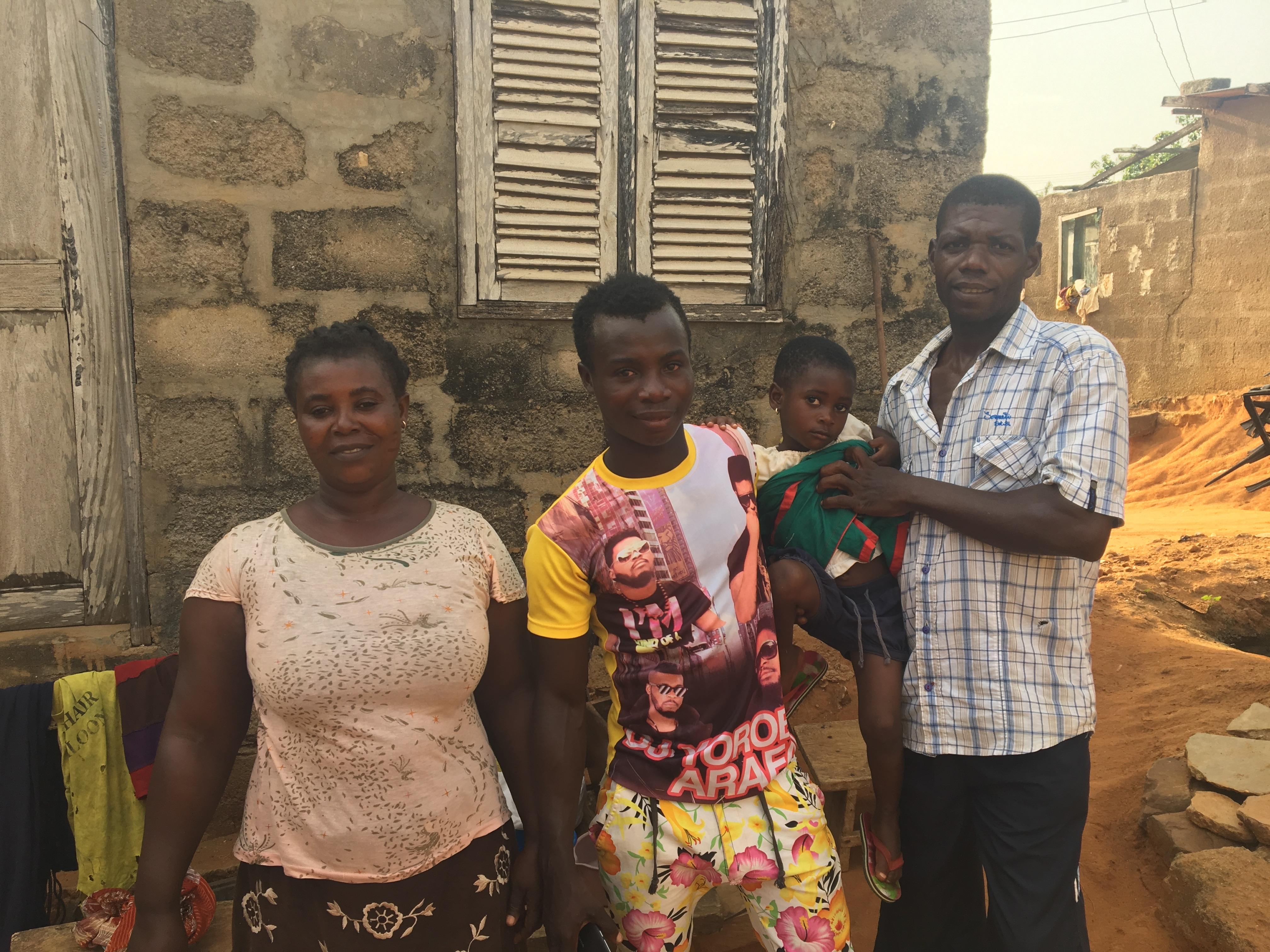 A family poses in front of a textured stone wall, illuminated by natural light. On the left, a woman with short, curly black hair wears a light-colored top with a floral pattern and a dark skirt. Next to her stands a young man with short hair, dressed in a colorful T-shirt featuring images of people, and patterned shorts. In his arms, he holds a small child wearing a green and red outfit. To the right, a man with short hair and a beard stands, wearing a button-up, plaid shirt and black trousers. Clothing lines are visible on the left side of the image, and the background features an outdoor space with dirt and another building. The overall scene conveys a warm family moment in a simple, rural setting.