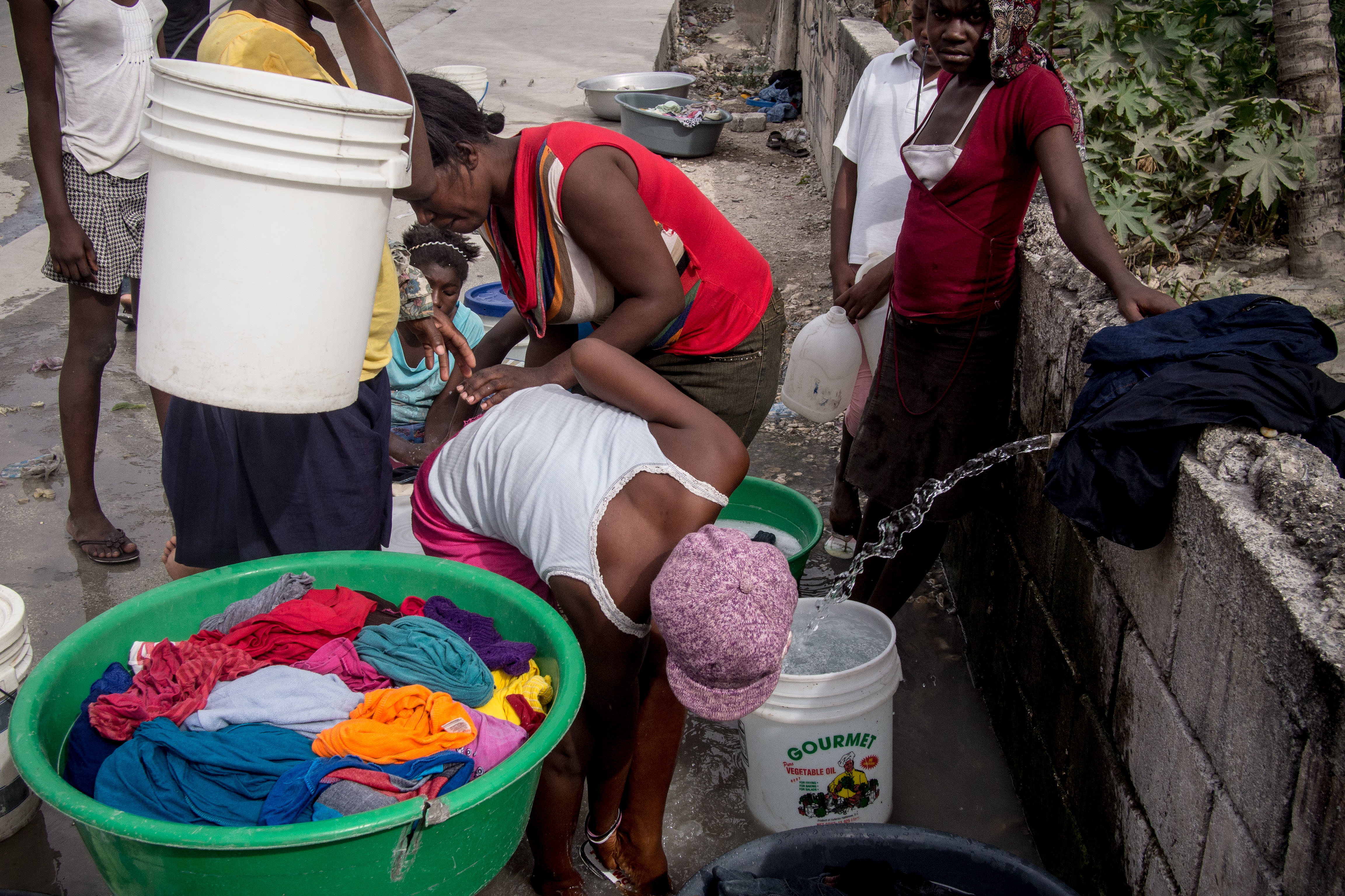 A group of women is engaged in washing clothes in a communal area. In the foreground, a large green basin filled with colorful, wet clothing is visible. One woman bends over the basin, scrubbing the garments. Nearby, another woman stands with a white bucket hanging from her arm, her clothing partially obscured. In the background, additional women are seen working together, some holding laundry items, while a stream of water flows from a nearby container into another bucket. The surroundings appear urban, with a concrete wall and scattered items on the ground, hinting at a lively community scene focused on the task of laundry.