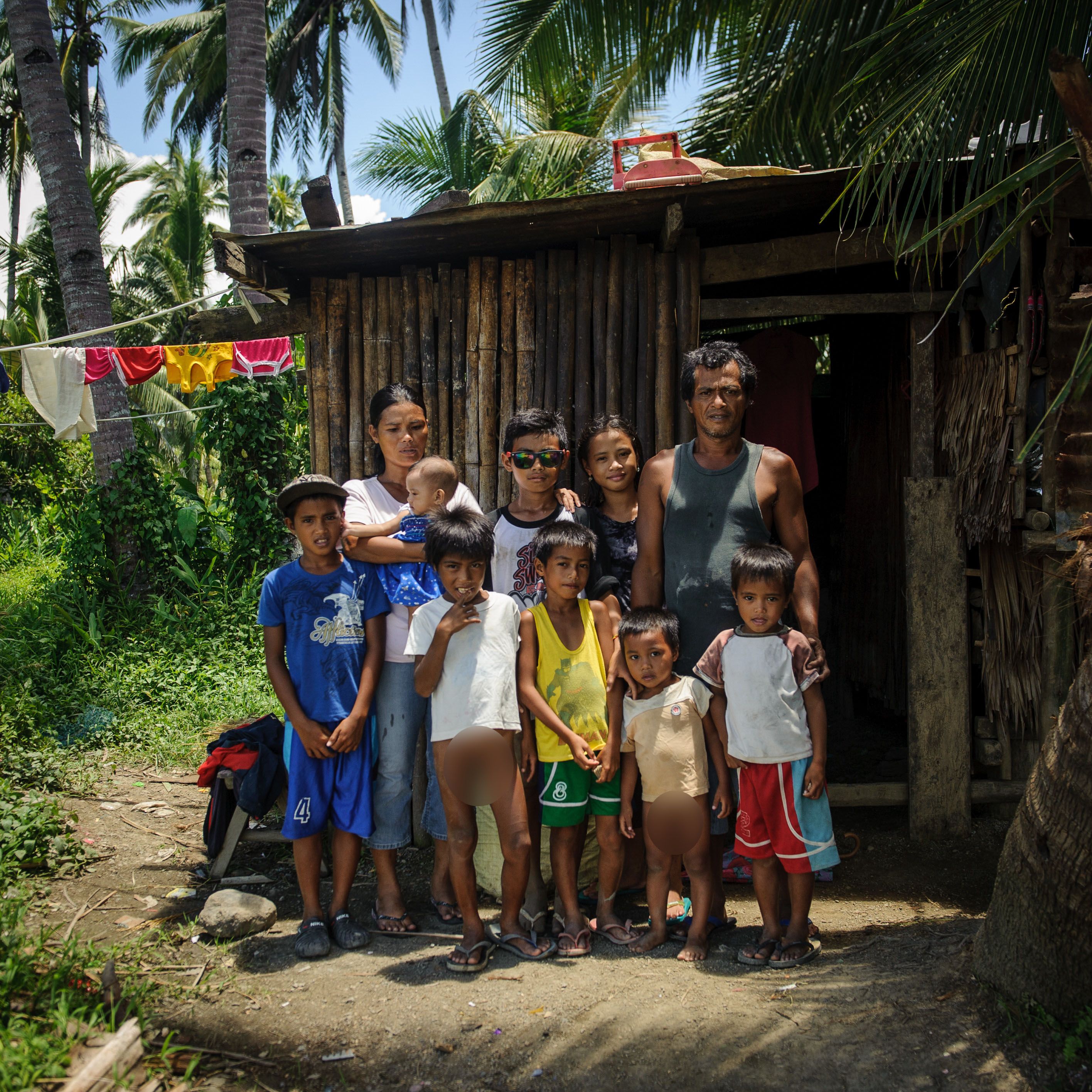 A group of thirteen people stands together outdoors in front of a rustic wooden house. The scene is vibrant and sunlit, with tall palm trees in the background. The group includes eight children, some holding hands, and a few adults. One woman cradles a baby. The children wear a variety of colorful clothing, with several in shorts and t-shirts, while the adults wear simple garments suitable for a warm climate. Laundry hangs to dry in the background on a clothesline. The atmosphere appears lively and familial, emphasizing community and togetherness.