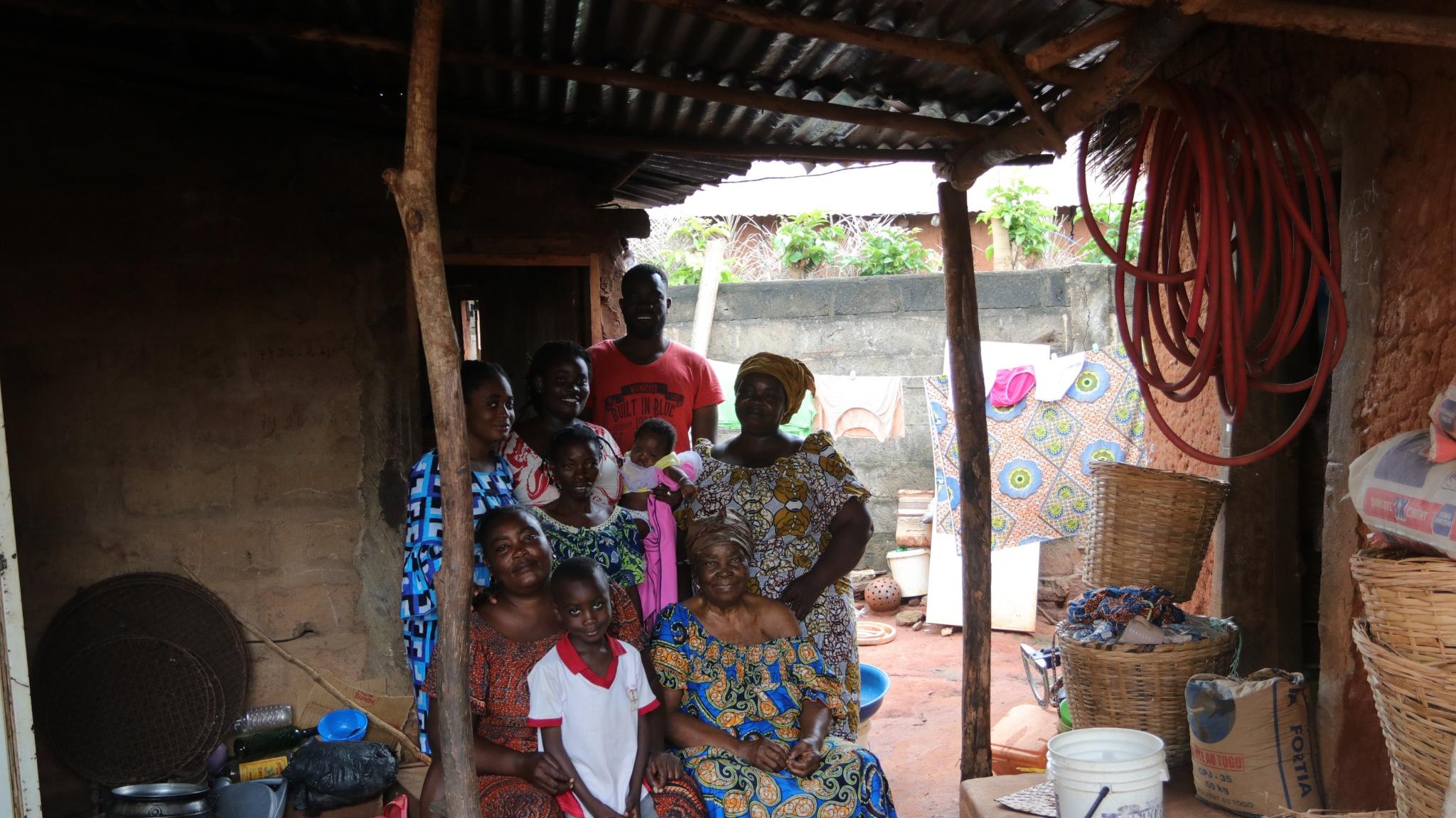 A group of nine people is gathered in a rustic setting under a corrugated metal roof. They are smiling and standing close together, showcasing a mix of traditional and casual clothing with vibrant patterns and colors. In the foreground, a woman is seated, wearing a blue and yellow patterned dress, while a young boy in a white shirt with red trim sits beside her. Behind them, others are positioned; a man in a red shirt stands at the back, holding a baby wrapped in a cloth. The background reveals a dirt floor with baskets, cooking utensils, and clothing hanging on a line. The overall atmosphere conveys warmth and community.