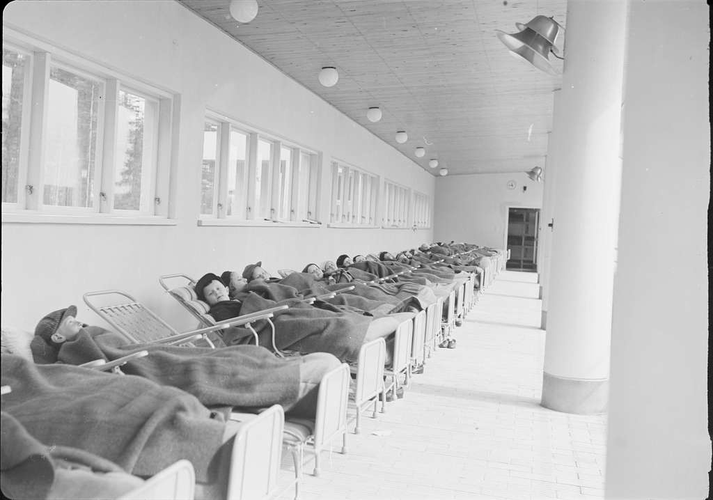 A long, narrow room lined with several beds occupied by children. Each child is wrapped in a blanket and reclining on a bed, with some wearing caps. Natural light streams in through multiple window panes along one side of the room. The walls are painted white, and the ceiling has wooden panels with light fixtures hanging down. The overall atmosphere appears calm and orderly, suggesting a space for rest or recuperation. A clock and a doorway can be seen at the far end of the room.
