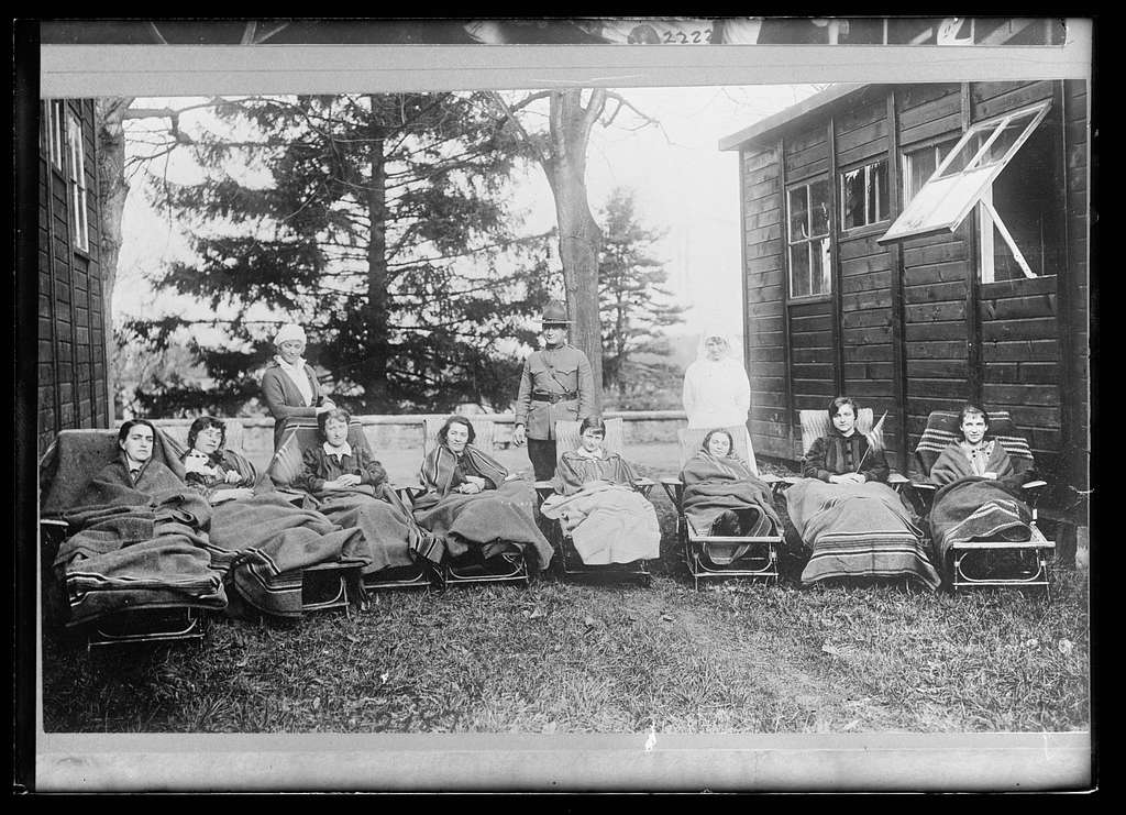 In a historical black-and-white photograph, a group of fourteen individuals is gathered outdoors. A man in a military uniform stands at the center, flanked by two women in white dresses, likely healthcare workers. The other eleven women are seated on lounge chairs, wrapped in blankets, suggesting a time of rest or recovery. In the background, there are tall trees and a wooden structure, likely a building or shelter, with windows. The scene conveys a sense of community and care, possibly in a context related to health or recovery during a past era. The overall atmosphere is calm and supportive, set in a natural environment.
